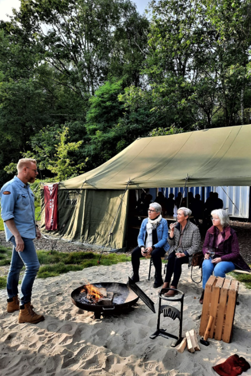 Foto van enkele personen rond een vuurkorf met op de achtergrond een legertent